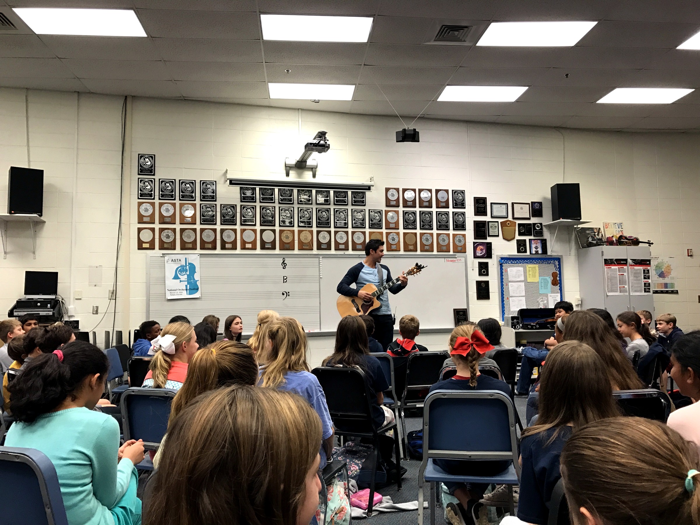 Chase d'Arnaud plays acoustic guitar for students in a school music classroom.