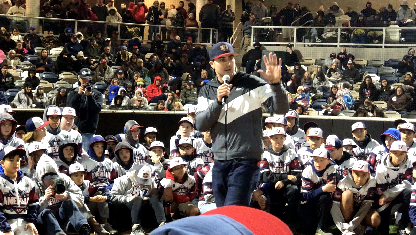 Chase d'Arnaud speaks into a microphone to young All-American players seated on the field at night.